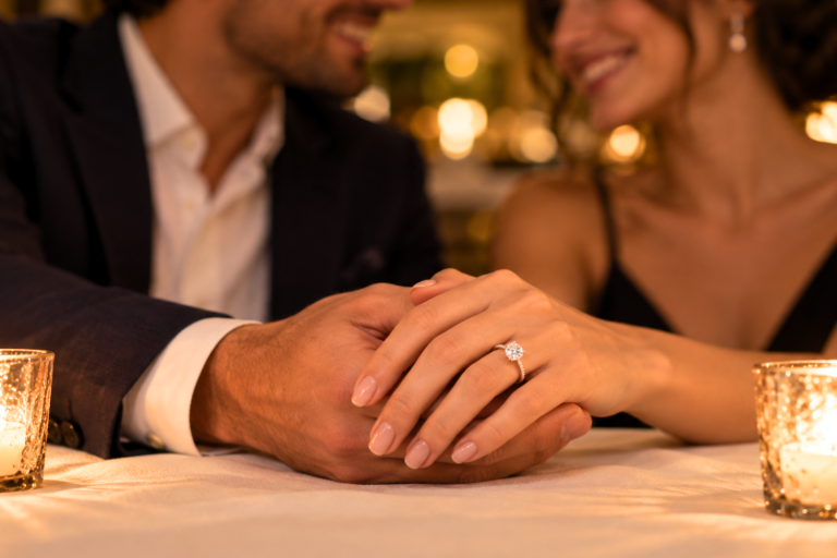 Newly engaged couple holding hands across a candlelit restaurant table, with the engagement ring in focus.