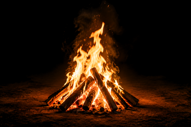 Bright wood bonfire burning on dark ground at night, with stacked logs, glowing embers, and orange flames.