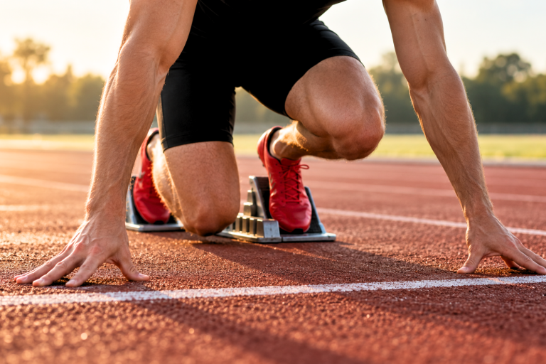 Runner crouches at the starting line with hands on the red track and bright red shoes braced against the starting blocks